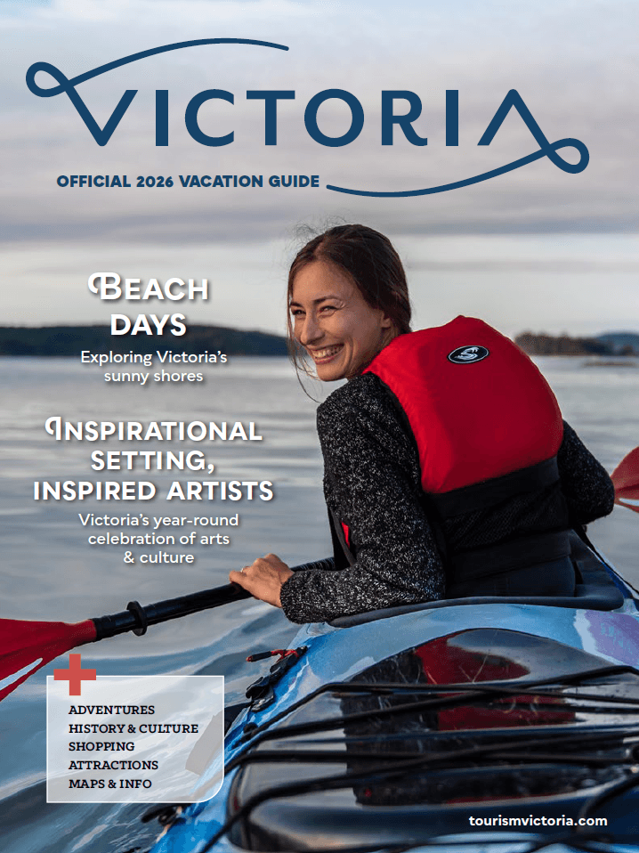 The cover of the 2026 Destination Greater Victoria Vacation Guide depicts a woman wearing a red life jacket and paddling a blue kayak looks back at her partner who is taking a photo of her.